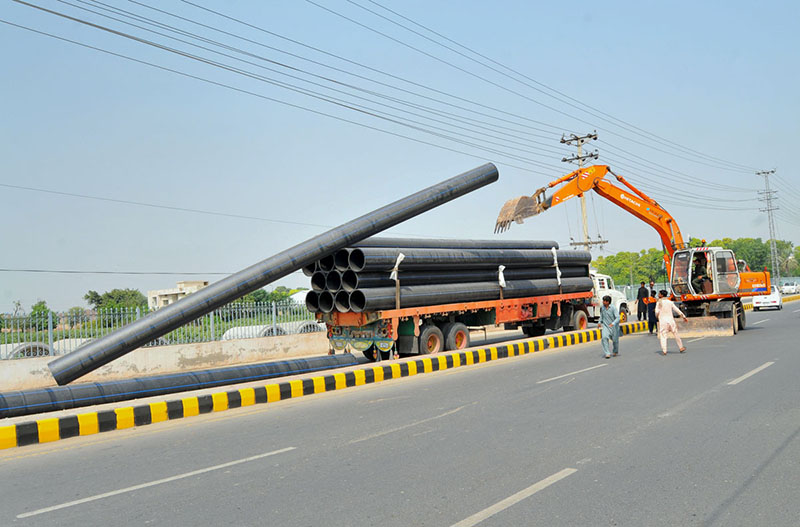 Labourers use an excavator to unload large plastic pipes from a truck along a road during ongoing development work