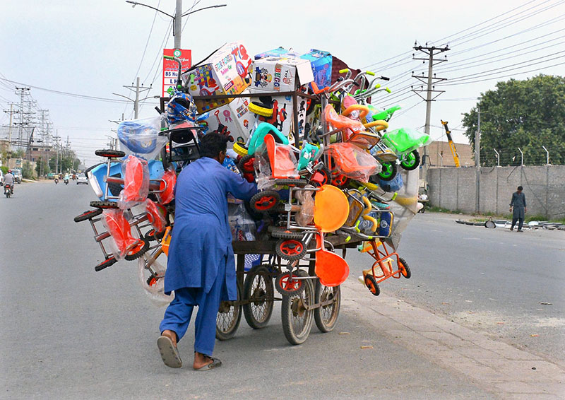 A vendor pushes a cart loaded with children's bicycles and other toys while crossing the road to earn a livelihood