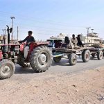 A tractor-trolley carrying a camel cart and other vehicles passes along the Site Area Road