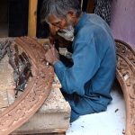 An elderly carpenter carves intricate designs into a wooden piece at his workplace