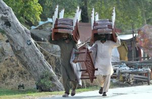 Street vendors carry wrapped plastic chairs and tables on their heads while walking along Vehari Road, navigating daily street trade to earn a livelihood.