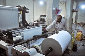 An operator monitors white fabric moving through a bag-making machine as workers produce eco-friendly non-woven bags at a local manufacturing facility.