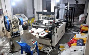 An operator monitors white fabric moving through a bag-making machine as workers produce eco-friendly non-woven bags at a local manufacturing facility.