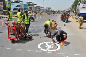Labourers paint road-safety markings for pedestrians near a busy intersection as part of ongoing traffic management and pedestrian safety measures.