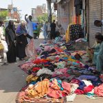 Vendors display colourful garments along a roadside to attract customers, while shoppers browse through the clothes at a busy market