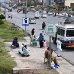 Commuters wait without any shelter at Pandora Bus Stop on IJP Road, exposed to harsh sun and rain, as the lack of proper covered waiting facilities at the busy bus stop continues to cause hardship for daily passengers and highlights the need for urgent attention from authorities