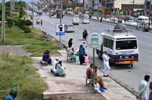 Commuters wait without any shelter at Pandora Bus Stop on IJP Road, exposed to harsh sun and rain, as the lack of proper covered waiting facilities at the busy bus stop continues to cause hardship for daily passengers and highlights the need for urgent attention from authorities