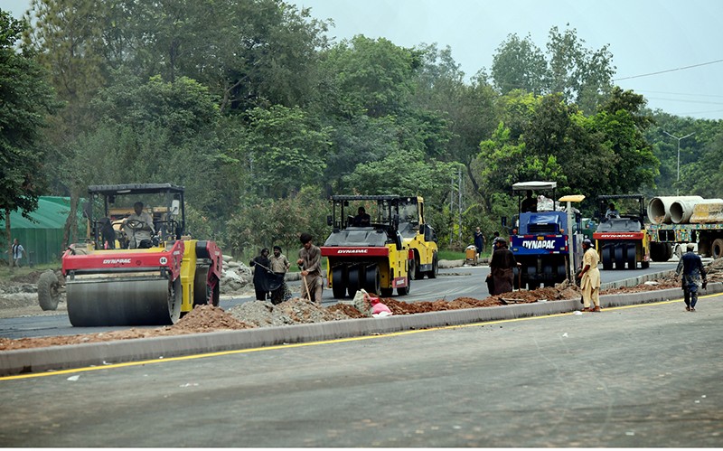 Heavy road rollers compact freshly laid asphalt at the Park Road Underpass project site, as construction machinery including Dynapac and Sakai rollers work alongside labourers to complete the carpeting of the road surface in the project's final phase