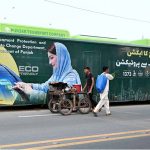 A street vendor wheels his handcart past a public bus displaying an environmental message from the Chief Minister of Punjab, part of a campaign urging citizens to protect nature and keep the province free from smog