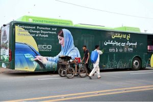 A street vendor wheels his handcart past a public bus displaying an environmental message from the Chief Minister of Punjab, part of a campaign urging citizens to protect nature and keep the province free from smog