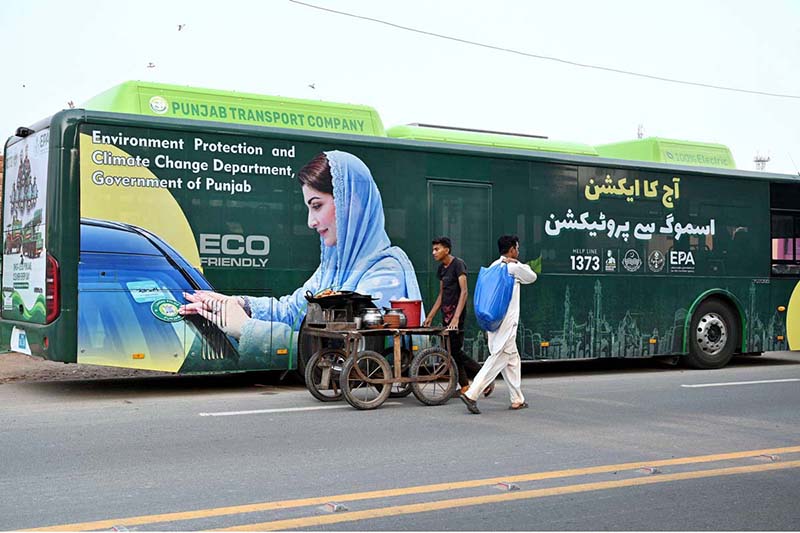 A street vendor wheels his handcart past a public bus displaying an environmental message from the Chief Minister of Punjab, part of a campaign urging citizens to protect nature and keep the province free from smog