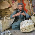 A man is crafting baskets, hand fans, and screens by hand from dried date palm leaves at his workplace