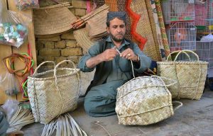 A man is crafting baskets, hand fans, and screens by hand from dried date palm leaves at his workplace