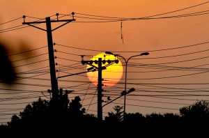 The setting sun creates a scenic silhouette of electricity transmission poles and wires against the evening sky.
