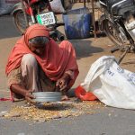 An elderly woman collects scattered grains from the roadside into a sack near a grain shop in the city