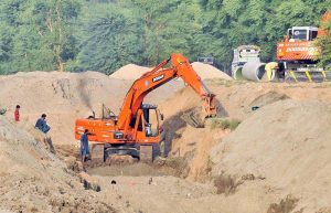 An excavator digs a deep trench as workers install a sewage pipeline along a roadside, with heavy machinery being used to excavate the ground for the ongoing development work.