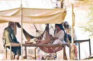 A nomadic woman weaves a traditional handmade basket from dry branches while sitting on a charpai under a makeshift canopy on the outskirts of the city.