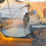 A worker boils sugarcane juice in a large pan while another tends the furnace fire to prepare traditional gur (jaggery) at a roadside setup in the city